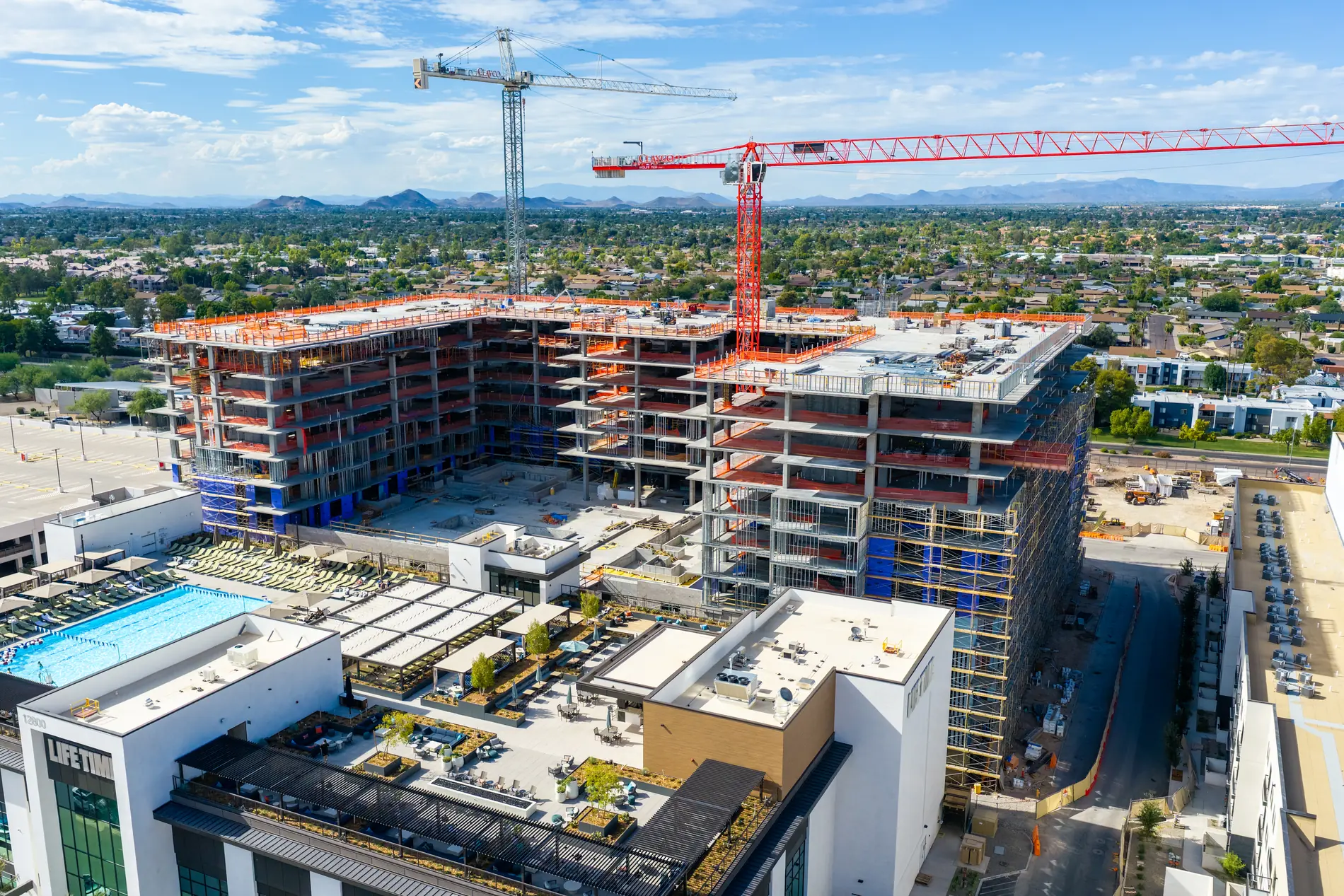 Mid-rise concrete structure under construction with two tower cranes, adjacent completed buildings and pool deck